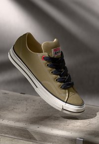 Light brown canvas sneaker with black laces, white rubber sole, and red accents. Features a textured upper and classic silhouette.