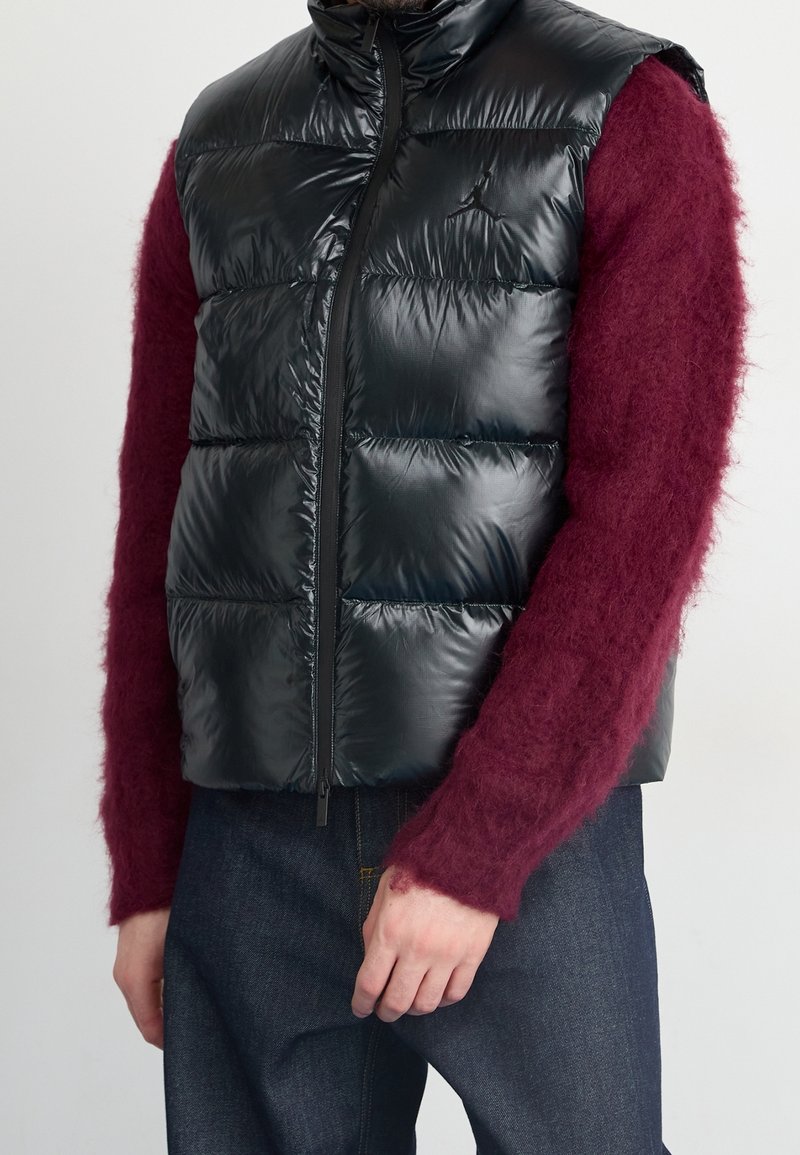 Person klädd i en glänsande svart vadderad väst över en burgunderröd fluffig tröja och mörkblå jeans, stående mot en enfärgad ljus bakgrund.