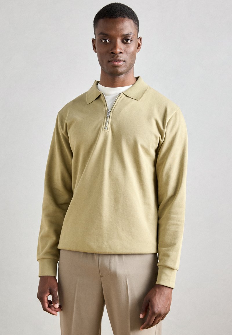 Világos olívazöld, póló stílusú, hosszú ujjú felső, negyed cipzárral, puha anyagból és klasszikus gallér kialakítással, fehér ing felett viselve.