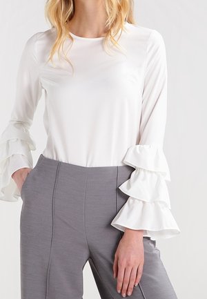 Femme portant un chemisier blanc à manches longues avec des volants en étages sur les manches et un pantalon gris taille haute, main reposant dans la poche.