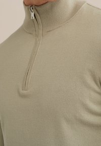 Närbild av en beige ribbad halvdraget tröjkrage och övre bröst på en person.