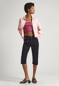 Mujer que lleva una chaqueta corta rosa sobre un top tubular fruncido en color malva, jeans negros hasta las rodillas y sandalias de tacón negras con tachuelas.
