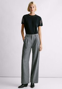 Camiseta negra de manga corta con detalle de flecos en los hombros y cuello con tachuelas, combinada con pantalones de pierna ancha grises plisados y botas de cuña negras.