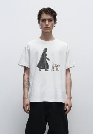Weißes Baumwoll-T-Shirt mit einem Grafikmotiv, das eine Figur in einem schwarzen Umhang zeigt, die einen beigen Roboter an einer grünen Leine führt. Kurze Ärmel, entspannter Schnitt.