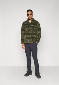 Pullover in pile verde scuro con motivi geometrici, collo con zip, abbinato a pantaloni scuri e stivali neri, in piedi contro uno sfondo neutro.