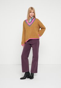 Pull en maille marron à col en V avec des accents roses, associé à un pantalon large violet foncé et des bottines noires.