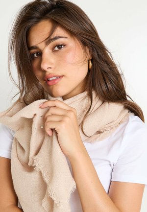 Écharpe beige en tissu tissé avec des bords frangés, une légère brillance et de petits embellissements en forme de cœur, drapée lâchement sur la main du porteur.