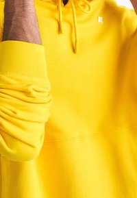 Homme portant un sweat à capuche jaune vif avec cordons tirés, bras droit levé, et manche légèrement remontée.