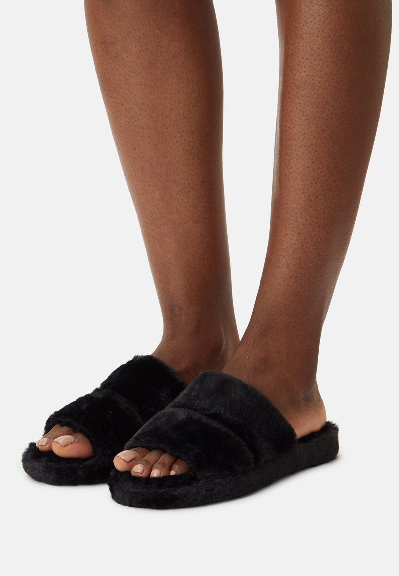 Pieds portant des chaussons noirs ouverts à bout ouvert, contre un fond clair uni, montrant les orteils et le bas des jambes.