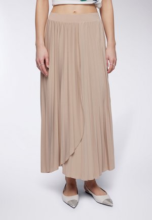 Gonna maxi plissettata beige con vita elasticizzata e orlo asimmetrico, realizzata in tessuto leggero. Abbinata a ballerine bianche a punta con borchie.