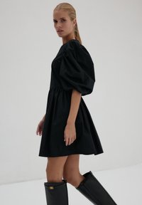 Kobieta z blond włosami związanymi w kucyk, ubrana w czarną sukienkę z bufiastymi rękawami i czarne wysokie kozaki, stoi na tle prostego jasnego tła.