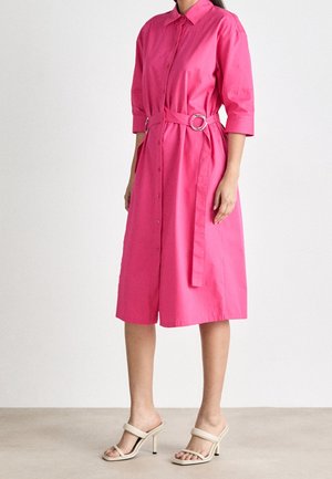 Femme portant une robe boutonnée rose vif longueur genoux avec manches trois-quarts et une ceinture assortie avec boucle argentée, associée à des sandales beiges à talons hauts.