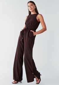 Brown sleeveless jumpsuit with a drawstring waist, wide legs, and side pockets; features gold hardware accents at the cuffs.