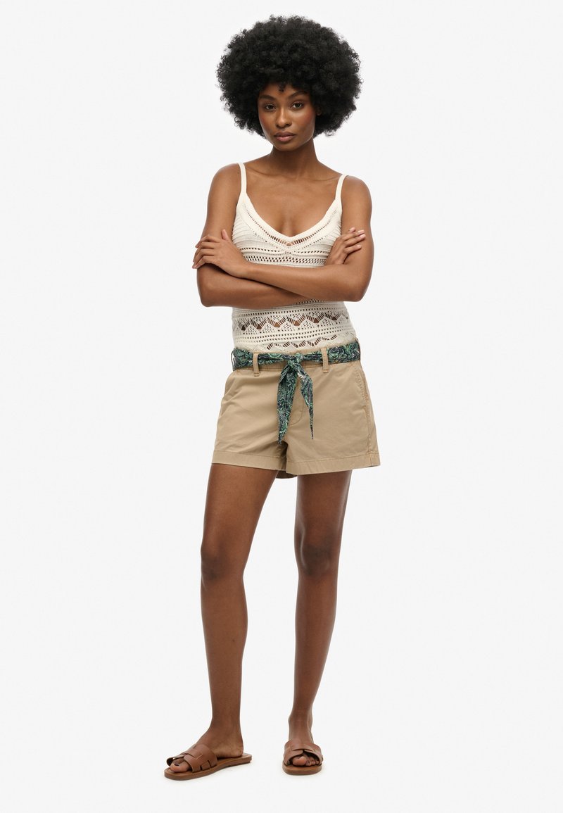 Beige shorts met een decoratieve groene riem met patroon, gecombineerd met een crème haakwerk tanktop. Model staat met de armen gekruist.