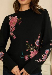 Melnā adīta džemperis ar krāsainiem ziedu izsādiem rozā, zilā un zaļā krāsā pāri pleciem un piedurknēm. Mīksts audums, klasiskā apaļā kakla izgriezumā.