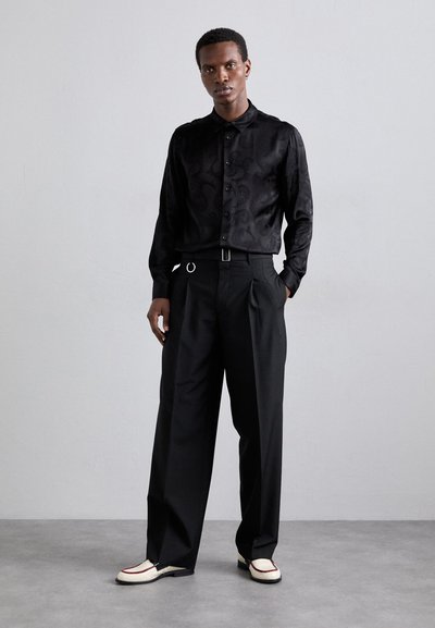 Camicia nera a maniche lunghe con un motivo sottile, pantaloni plissettati neri e scarpe bianche con dettagli neri. Modello in piedi su uno sfondo grigio.