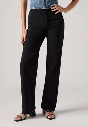 Femme portant un pantalon noir ample taille haute, un gilet court en denim et des sandales noires à talon bloc et bout ouvert, debout avec une main sur la hanche.