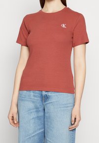 Ribbed, krátký rukáv tričko v hnědočervené barvě s kulatým výstřihem a malým bílým logem CK na levé hrudi. V kombinaci se světle modrými džíny.