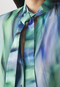 Blusa di seta blu e verde con dettaglio al collo a cravatta. Presenta un motivo a onde sottile, una consistenza morbida e una chiusura con bottone al colletto.