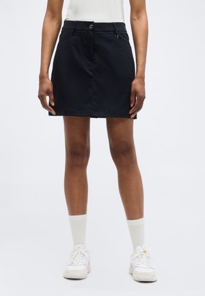 Woman wearing a black skirt, white socks, white sneakers, and a white top, standing against a plain light background.