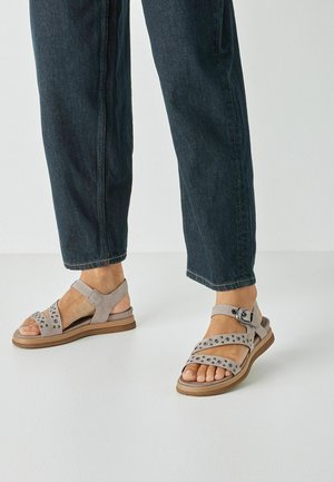 Gray suede sandals with adjustable straps, metallic studs, and a low wooden sole. Worn with dark denim cropped pants.