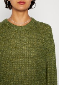 Grön stickad tröja med löst, strukturerat väv och rundad halsringning. Materialet har en subtil glans och variationer i gröna nyanser.