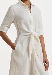 Light beige linen shirt dress with short sleeves, a button-down front, and a tied waist detail. Features side pockets and a relaxed fit.