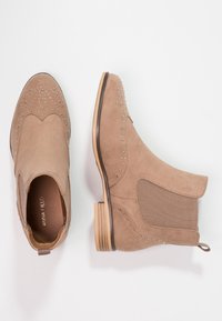 Beige nubuck nilkkurit koristeellisilla niiteillä, joustavilla sivupaneeleilla ja puukorolla. Sileä pinta ja pyöristetty kärki.