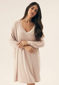 Jasnoróżowa sukienka z długim rękawem, z dekoltem w kształcie litery V i luźnym krojem. Gładki materiał, do kolan, z bocznymi rozcięciami dla wygody ruchu.