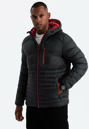 Doudoune noire avec des accents rouges, dotée d'une capuche, de poches zippées et d'une texture matelassée. Portée par-dessus une chemise blanche.