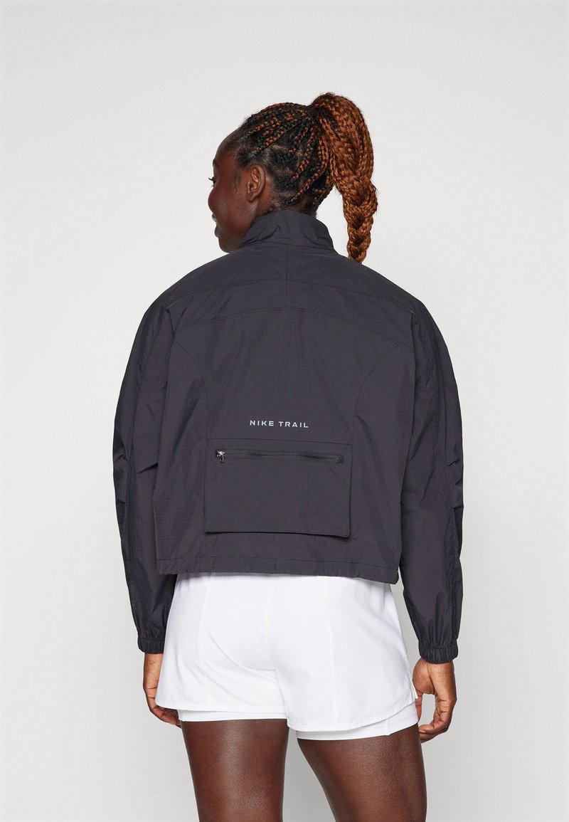 Schwarze, leichte Nike Trail-Jacke mit hohem Kragen, elastischen Bündchen und einer Reißverschlusstasche auf der Rückseite. Hat ein minimalistisches Design ohne Muster.