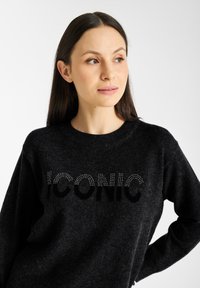 Zwarte trui met een ronde halslijn, voorzien van het woord "ICONIC" in fonkelende studs, gemaakt van een zachte, gestructureerde stof.