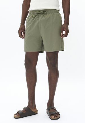 Olivgrüne Badehose mit elastischem Bund und Kordelzug. Der Stoff wirkt leicht und glatt. Getragen mit dunkelbraunen Sandalen.
