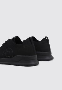 Zapatillas deportivas negras hechas de material de punto texturizado. Cuentan con una punta redondeada, suela plana y un cuello bajo, con un logo en el costado.
