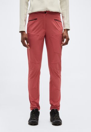 Osoba oblečená v koralovo ružových vonkajších nohaviciach s čiernymi vreckami na zips a čiernych turistických topánkach, stojaca na bielom pozadí.