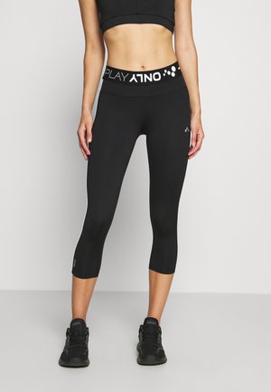 Vrouw draagt zwarte cropped legging met "ONLY PLAY" gebrande tailleband en zwarte sportschoenen, staand tegen een effen achtergrond.