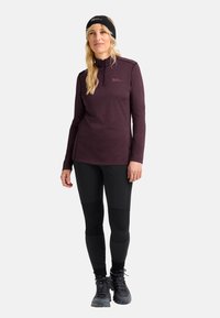 Donna che indossa una felpa zip-up bordeaux, leggings neri, scarpe da trekking e un cerchietto nero, in piedi su uno sfondo bianco uniforme.