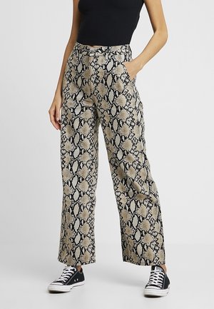 Femme portant un pantalon large taille haute avec un motif peau de serpent beige et noir, un haut sans manches noir et des baskets noires avec des semelles blanches.