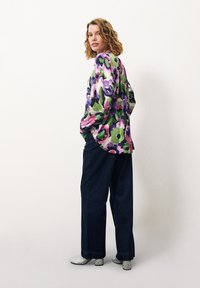 Bluse mit floralem Muster in Grün, Pink und Lila; lockere Passform und lange Ärmel. Kombiniert mit weit geschnittenen, dunklen Jeans und glitzernden Blockabsätzen.