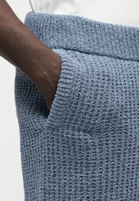 Main à la peau foncée partiellement dans la poche d’un pantalon en maille bleu clair texturé porté avec un haut blanc.