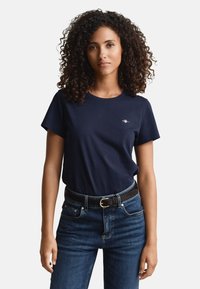 Navy cotton t-shirt with short sleeves, round neckline, and small embroidered logo on the chest, paired with dark denim jeans and a black belt.