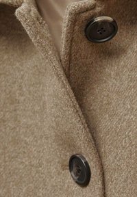 Manteau en laine marron avec une finition texturée, doté d'un col à revers et de deux boutons noirs. Gros plan sur les détails des boutons et le bord du col.