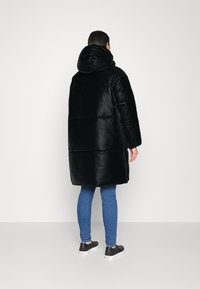 Cappotto piumino nero con cappuccio, caratterizzato da un design trapuntato. Indossato sopra jeans blu e sneakers nere. Texture liscia, vestibilità di lunghezza media.