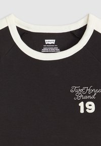 Černé tričko s krémovým límcem a raglánovými rukávy. Na přední straně jsou bíle vyšívané nápisy "Two Horse Brand" a "19". Materiál: bavlna.