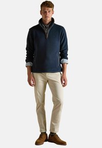 Mladý muž oblečený v námorníckom fleecovom svetríku na zips, pruhovanej košeli s vyhrnutými rukávmi, béžových nohaviciach a hnedých semišových topánkach, stojí čelom.