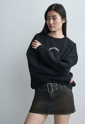 Mlada ženska, oblečena v črno široko majico Palm Angels in črno mini krilo iz jeansa s širokim pasom, pozira s prekrižanimi rokami in gleda vstran.