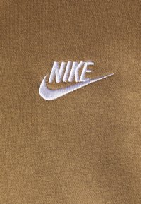 Logo Nike ricamato in bianco su un tessuto texturizzato di colore marrone dorato. Il design presenta uno stile di branding sportivo classico.