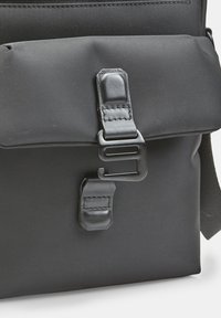 Borsa a tracolla nera in tessuto caratterizzata da una superficie liscia, una chiusura a pattina e una fibbia in plastica. Include un dettaglio di cucitura sulla pattina.