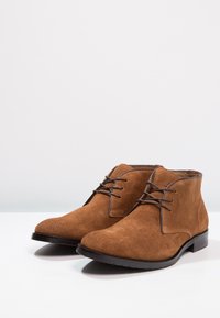 Bottes courtes en daim marron avec un bout rond, un système de laçage, et une semelle en caoutchouc noire. Texture lisse avec des coutures décoratives contrastantes.