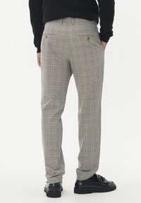 Mężczyzna w szarych spodniaczych w kratę i czarnych mokasynach, stojący z rękami w kieszeniach, częściowo widoczna czarna góra.